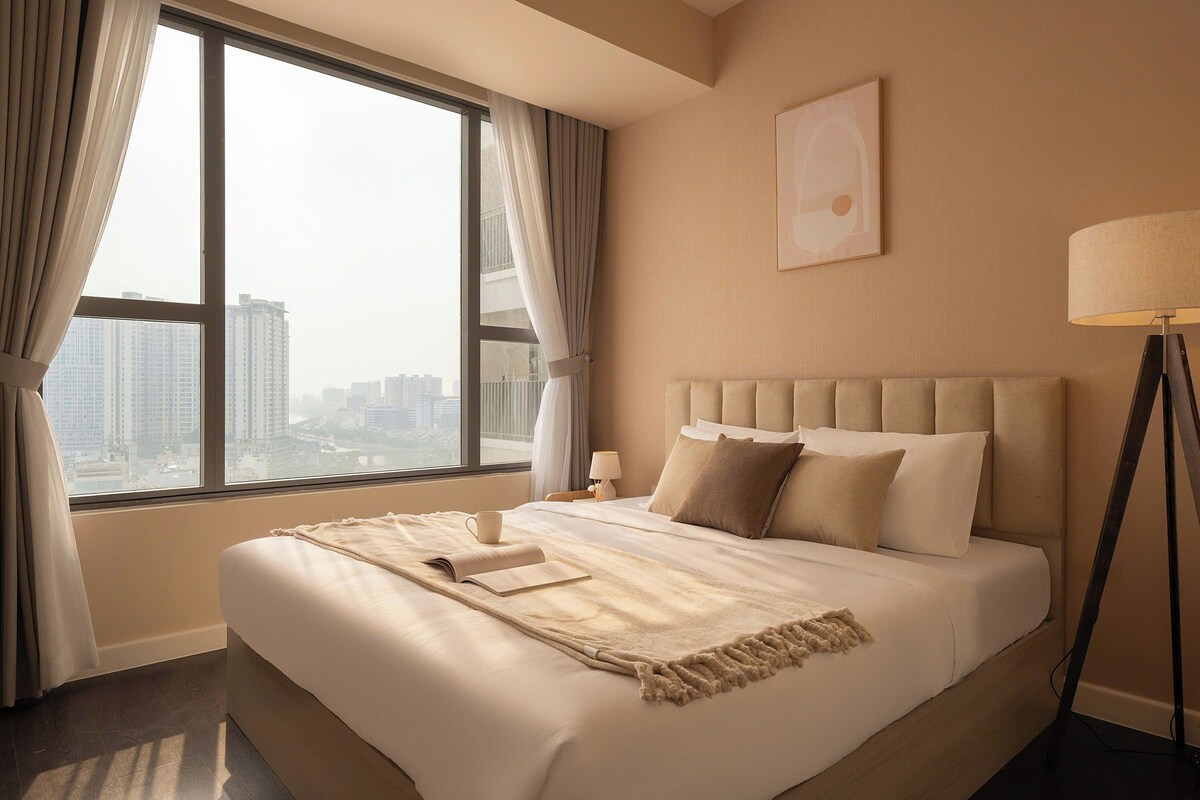 A hotel room with a large bed, a bedside table, a lamp, and a view of the city through the window.