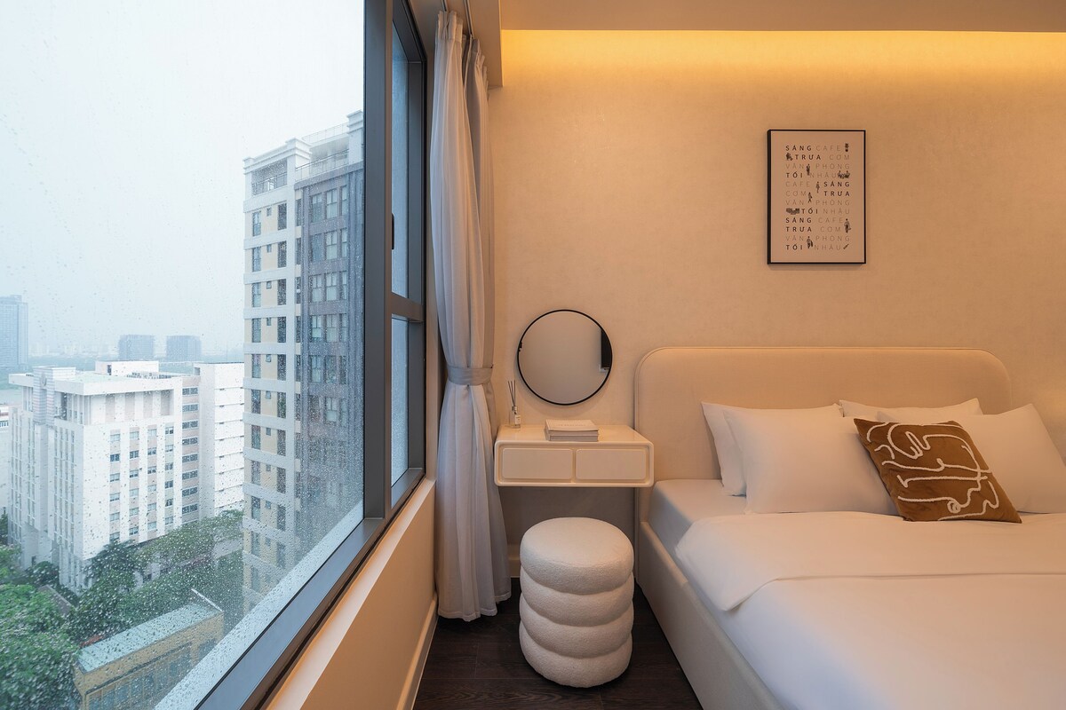 A hotel room with a large window, a bedside table with a mirror, a round stool, and a bed with pillows.