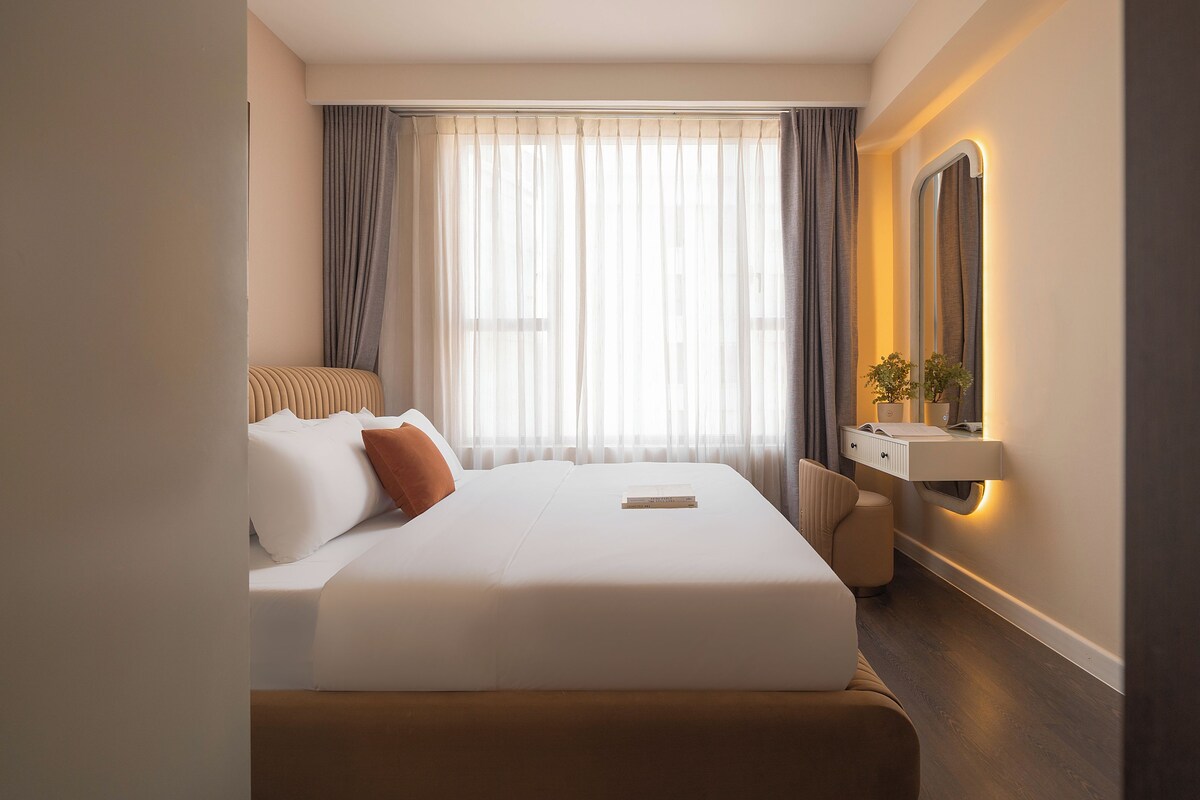 A hotel room with a bed, a bedside table with a mirror, a small desk, and a window with curtains.