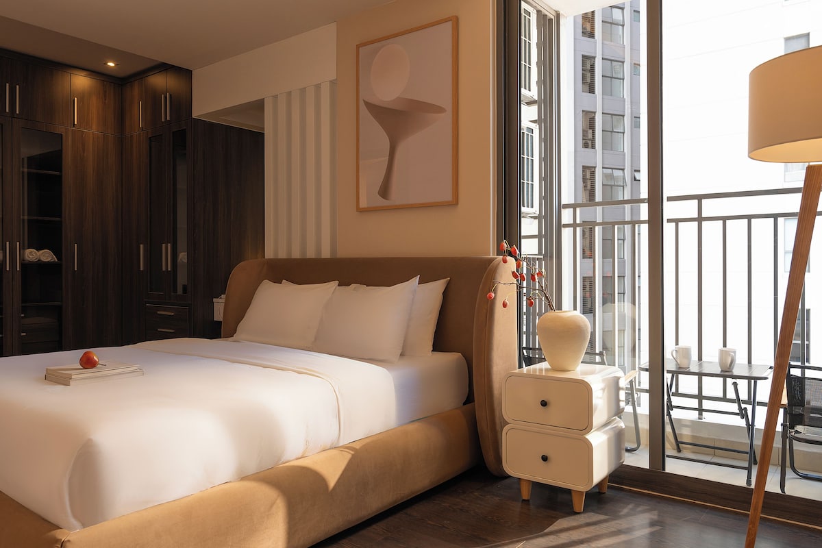A hotel room with a large bed, a bedside table, a nightstand, a window with a view of buildings, and a framed artwork on the wall.