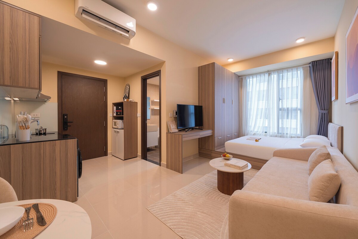 A modern hotel room with a kitchenette, a flat-screen TV, a sofa, and a large window with curtains.