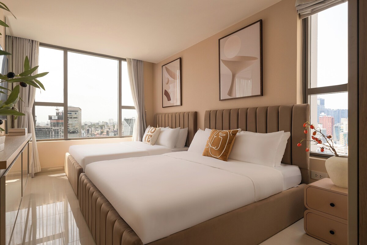 A hotel room with two beds, a desk, and a large window offering a city view.
