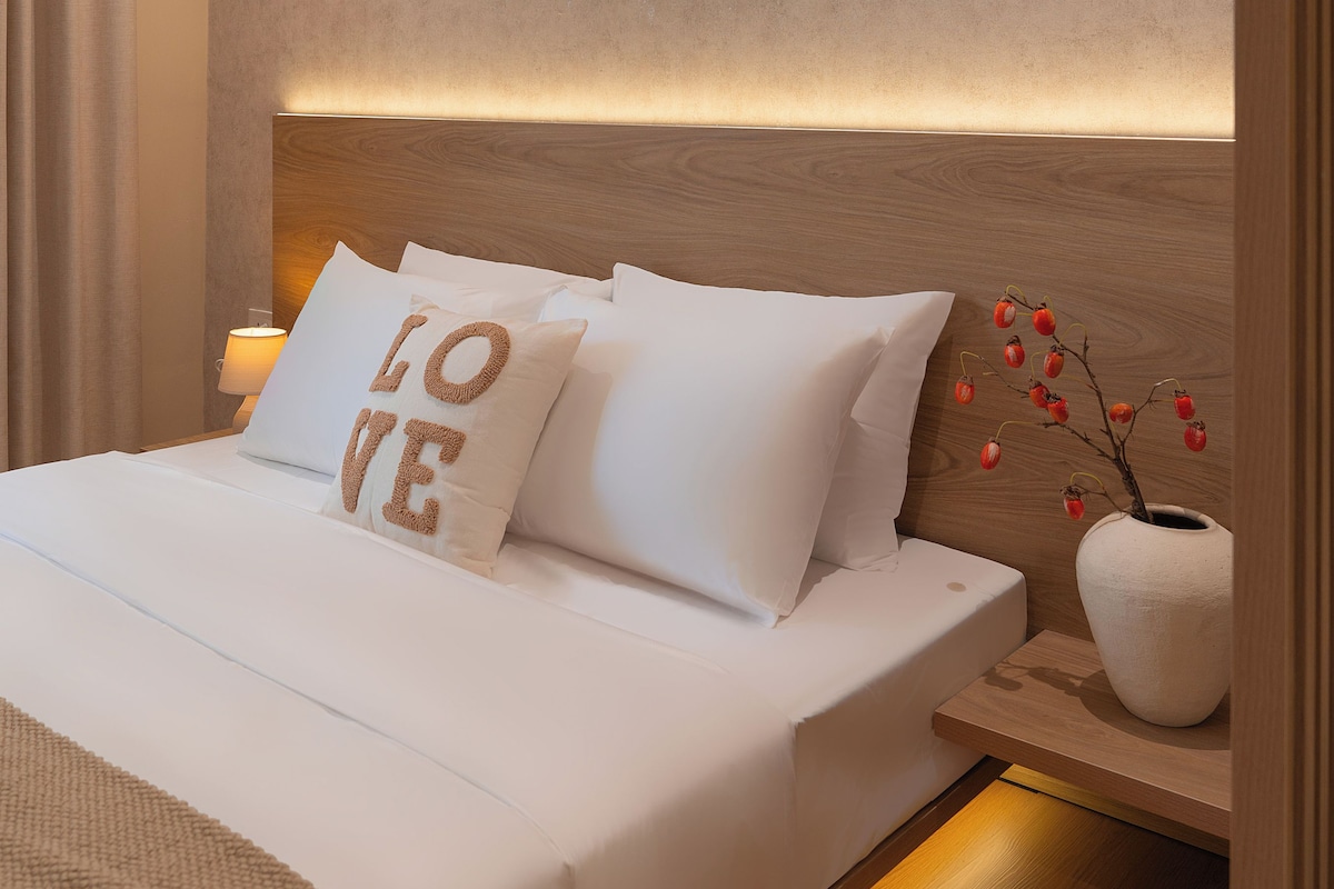 A modern hotel room with a large bed, a headboard with integrated lighting, a bedside table with a lamp, and a decorative wall piece.