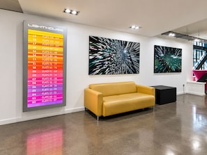 Ein modernes Interieur mit einem gelben Sofa, abstrakter Kunst an den Wänden und einem beleuchteten Schild mit Zimmernummern und -typen.