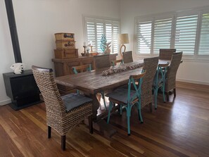 Een eetgedeelte met een houten tafel en rieten stoelen, een open haard en uitzicht door witte luiken.