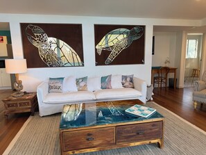 A living room with a white sofa, a glass-top coffee table, and two wall-mounted turtle paintings. - Relaxing getaway with the family  on the water. (South Stradbroke)