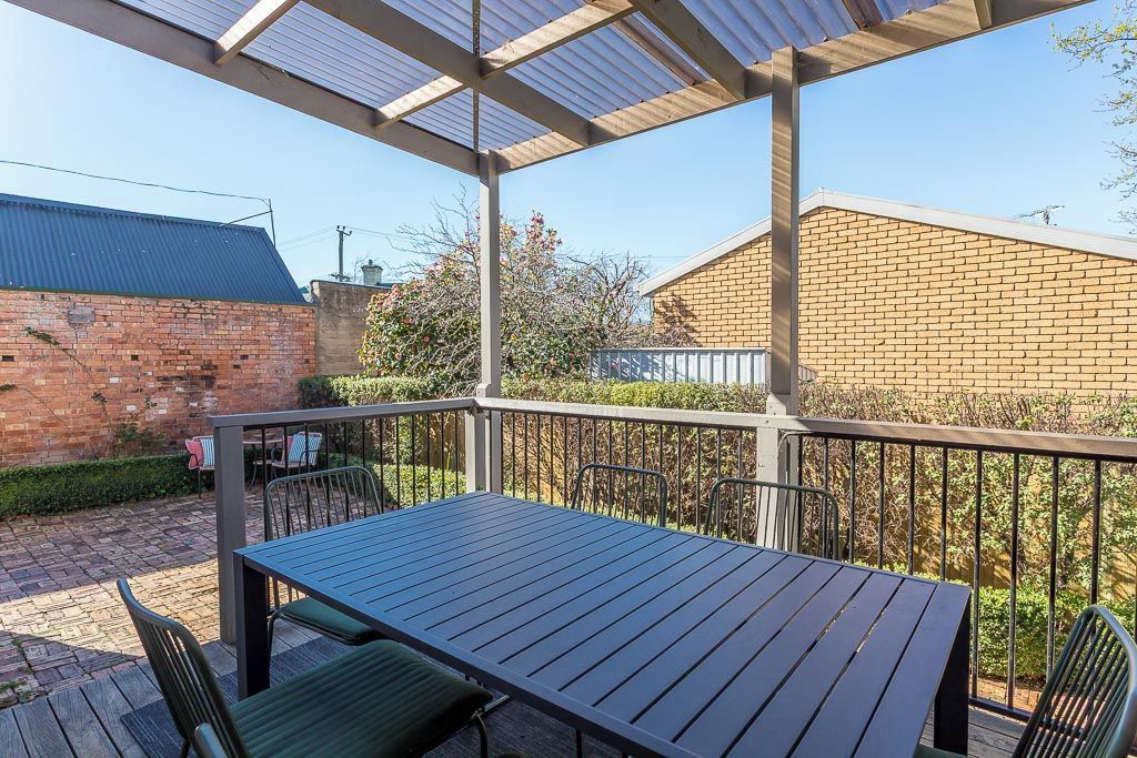 A covered outdoor patio with a table and chairs, surrounded by a metal railing and a brick wall.