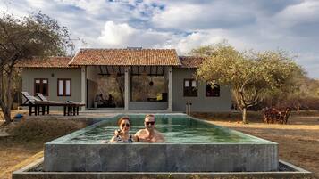 A swimming pool with two people in it, a covered outdoor seating area, and a building with a tiled roof.