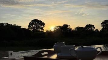 A wooden table with a tray holding two white cups and a teapot, set against a sunset backdrop with trees and a clear sky.