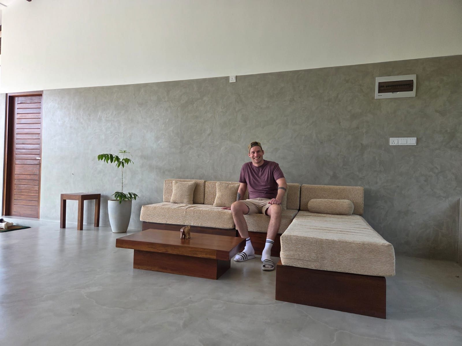 Ein Mann sitzt auf einem Sofa in einem modernen Wohnzimmer mit einer Betonwand, einem kleinen Tisch und einer Topfpflanze.