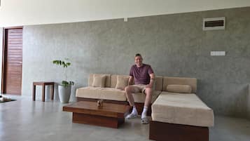 Ein Mann sitzt auf einem Sofa in einem modernen Wohnzimmer mit einer Betonwand, einem kleinen Tisch und einer Topfpflanze.