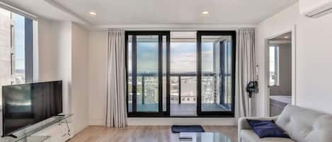A modern living room with a flat-screen TV, a glass coffee table, a sofa, and large sliding glass doors offering a city view.