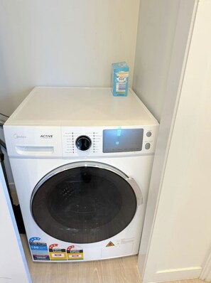 Een compacte, witte wasmachine met een digitaal display en bedieningspaneel, geplaatst in een kleine wasruimte met een houten vloer.