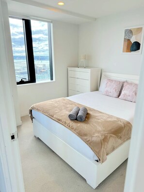 A modern bedroom with a large bed, a nightstand, a dresser, and a window with a city view. - Stunning Seaview+Skytower View 2 Bedroom Apartment (Auckland central)