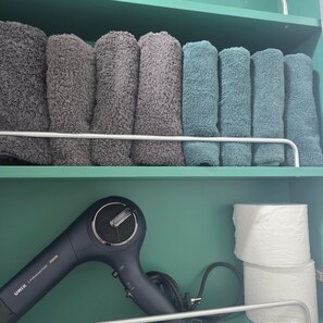 A shelf with neatly folded towels in various shades of gray and blue. Below, there is a black hairdryer with a cord and a roll of white paper towels. - [5 Min to Myeongdong] Twin Beds · Walk-in Closet · Separate Shower (Seoul)