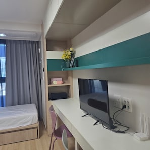 Una habitación de hotel con una cama, un escritorio con una computadora, una silla y una estantería con un jarrón de flores.