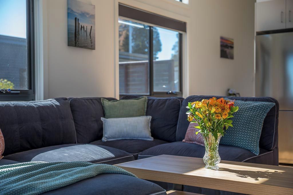 Uma sala de estar moderna com um sofá de cor escura, uma mesinha com um vaso de flores e uma janela grande que proporciona iluminação natural.