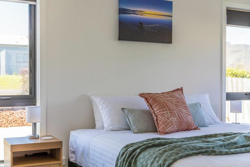 Een slaapkamer met een bed, kussens, een nachtkastje, een lamp en een schilderij aan de muur.