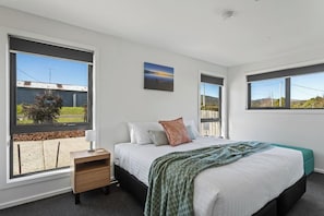 Een slaapkamer met een groot bed, een nachtkastje, een raam met uitzicht op een gebouw en groen, en een schilderij aan de muur.