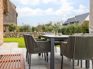 An outdoor patio with a table and chairs, a stone wall, and a grassy area. - Bals Westerlandstr. 11b Lee Reethus (Wenningstedt-Braderup)