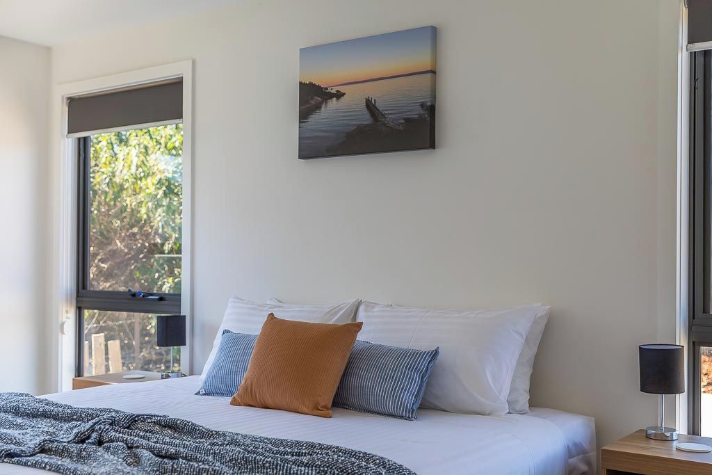 A bedroom with a bed, pillows, a blanket, a painting, a nightstand, a lamp, and a window with a view of trees.