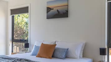 A bedroom with a bed, pillows, a blanket, a painting, a nightstand, a lamp, and a window with a view of trees.