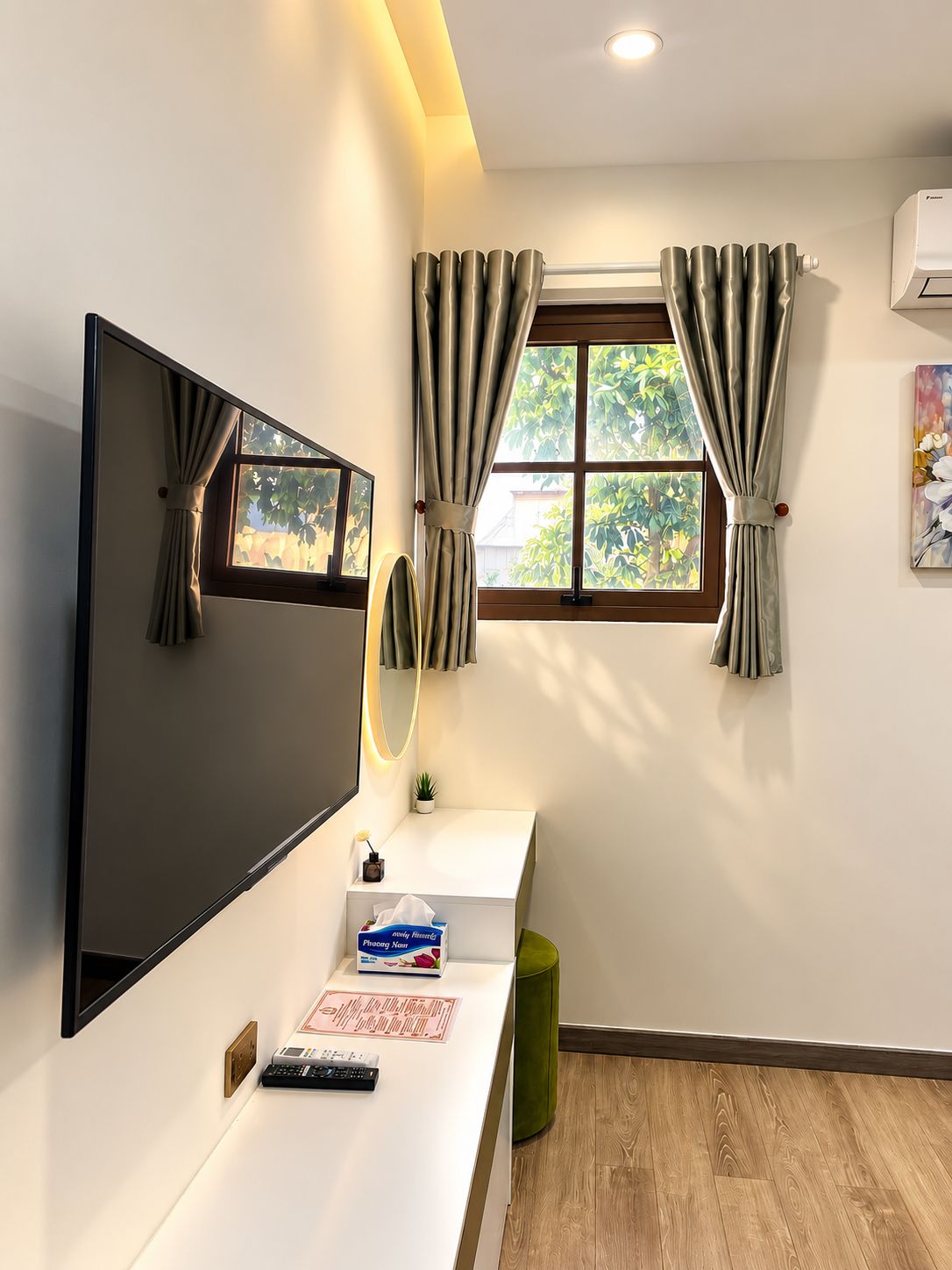 Een hotelkamer met een flatscreen televisie aan de muur, een klein nachtkastje met een afstandsbediening, een spiegel en een raam met gordijnen.