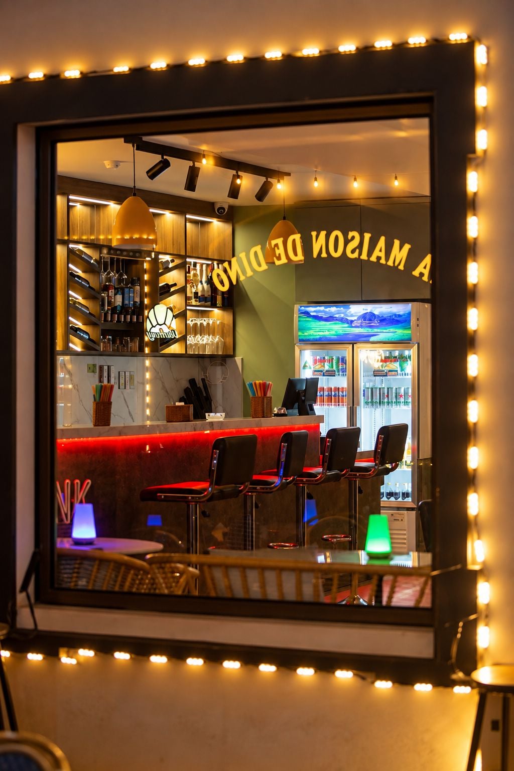 A bar area with a red counter, multiple chairs, shelves with bottles, and a neon sign.