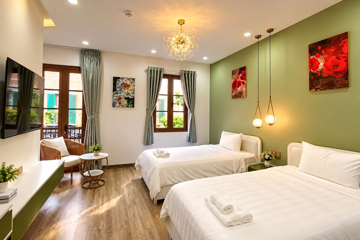 A hotel room with two beds, a desk with a chair, a small table with a plant, and a view of greenery outside.