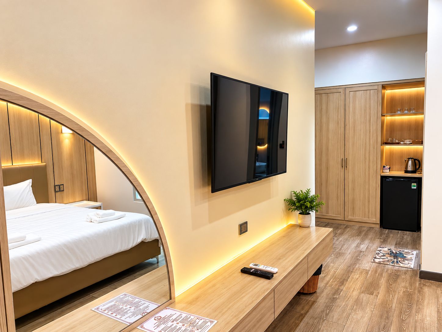 A modern hotel room with a bed, a flat-screen TV mounted on the wall, a small potted plant, a wooden bench, and a kitchenette area.