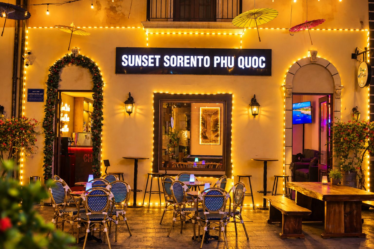 Un área de estar al aire libre con mesas y sillas, un letrero que dice 'SUNSET SORENTO PHU QUOC' y luces decorativas.