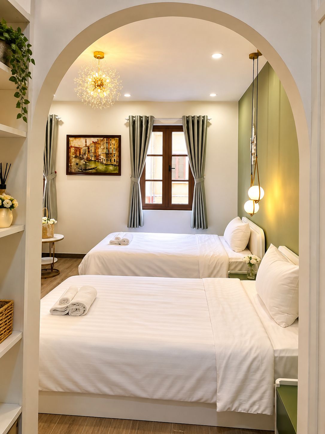 A hotel room with two beds, a painting of a cityscape, a window with curtains, and a chandelier.