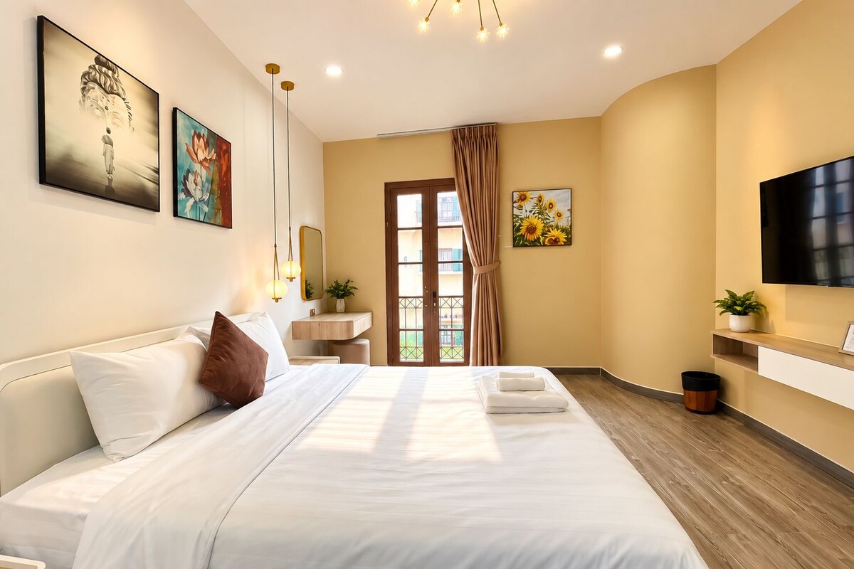 A modern hotel room with a large bed, a flat-screen TV, a bedside table, a mirror, and artwork on the walls.