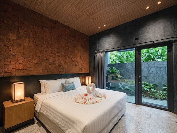 A modern bedroom with a brick wall, a large bed with white linens, a nightstand, and a view of a garden through a sliding door.