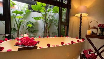 A bathtub with rose petals and a candle, a lamp, and a basket of roses.
