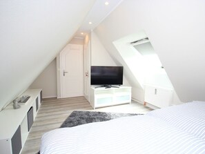 A bedroom with a sloped ceiling, a television on a white dresser, a door with a red handle, and a window. - Holiday Apartment With 2 Bedrooms (Sylt)
