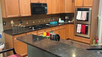 Una cocina con armarios de madera, un microondas negro, un refrigerador de acero inoxidable, una encimera de granito y un taburete de bar.