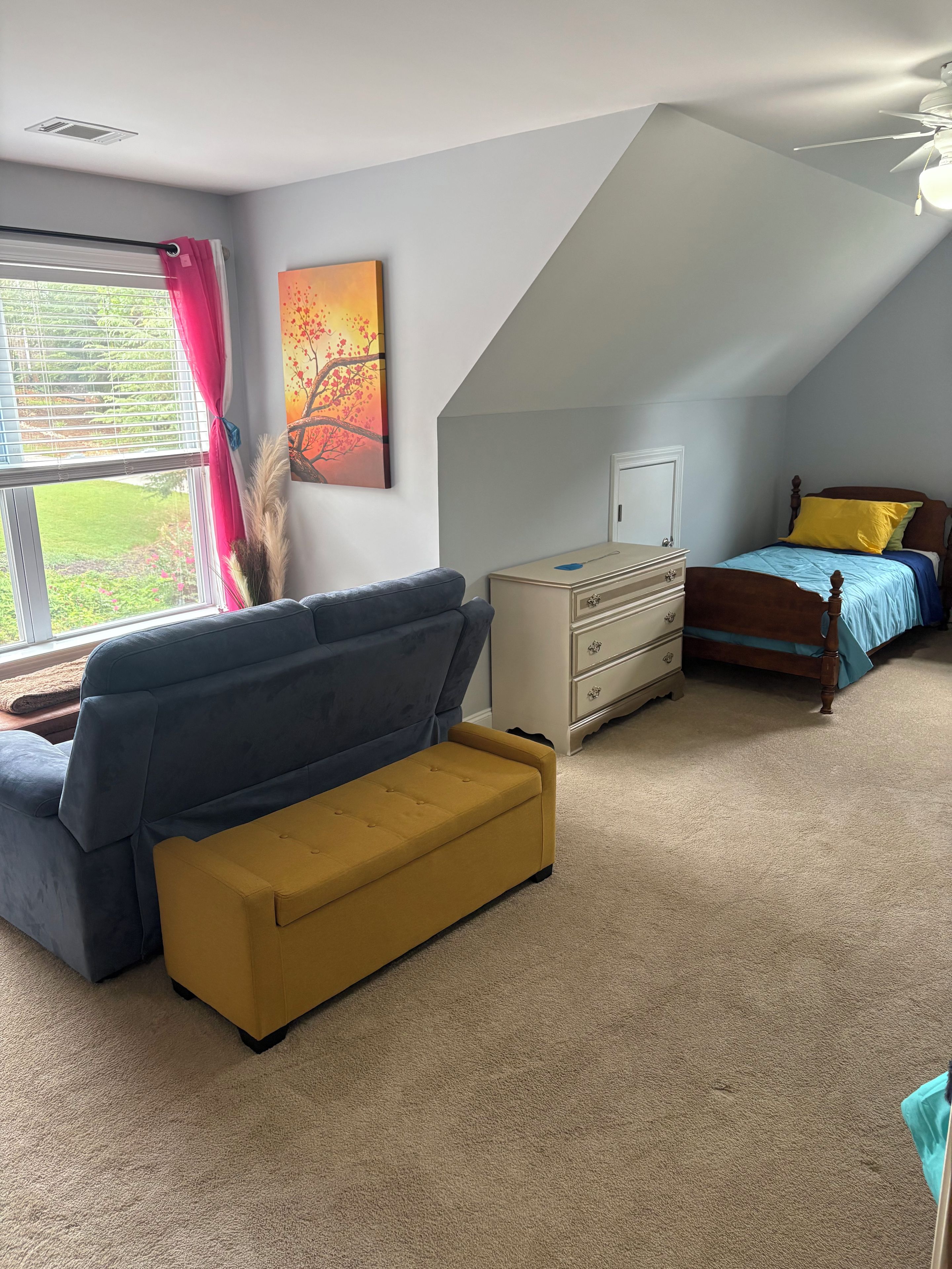 A bedroom with a sloped ceiling, a bed with a blue and yellow bedspread, a blue sofa, a yellow ottoman, a dresser, and a window with curtains.