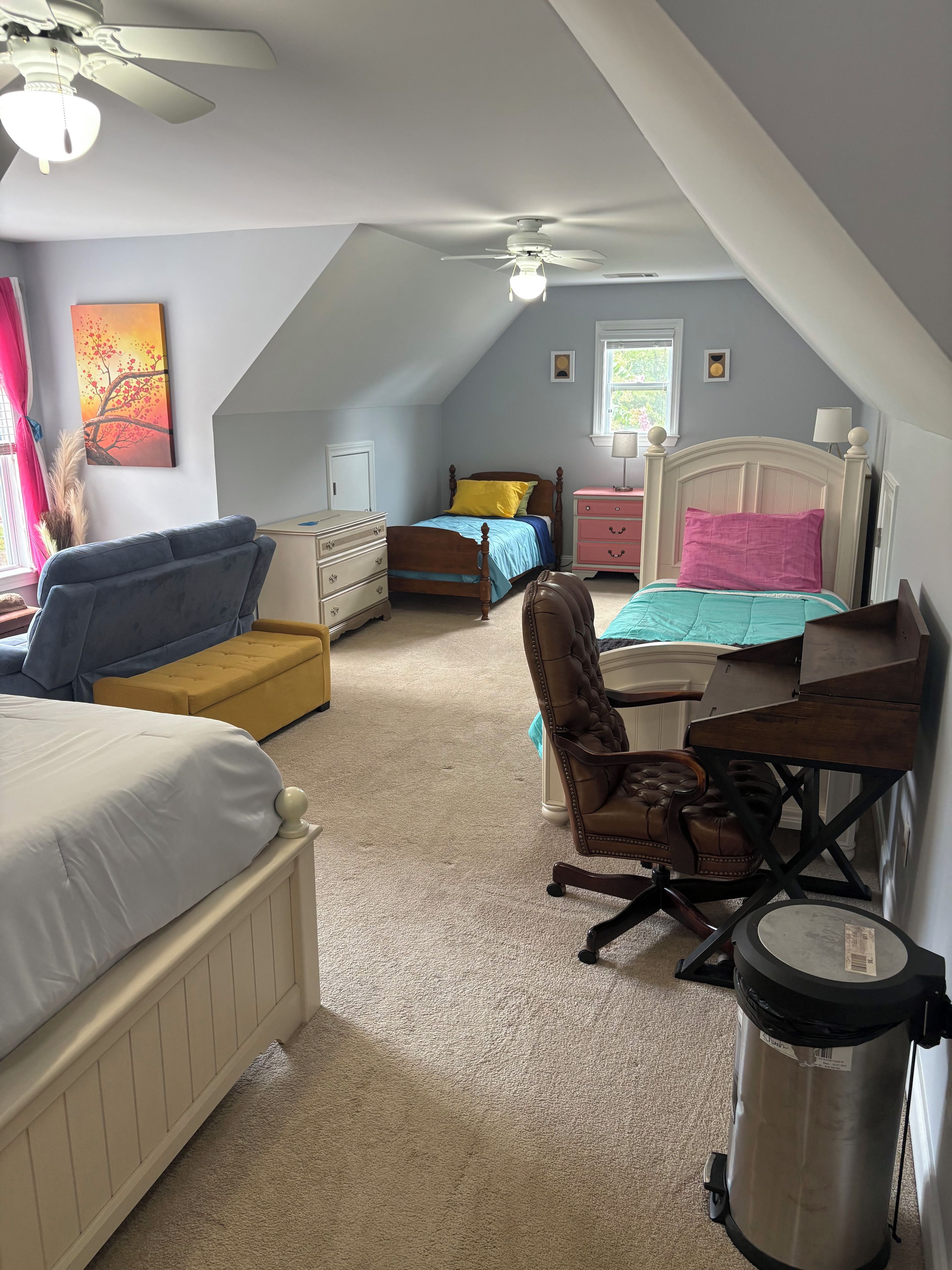 A bedroom with a sloped ceiling, a bed, a desk, a chair, a dresser, and a trash can.