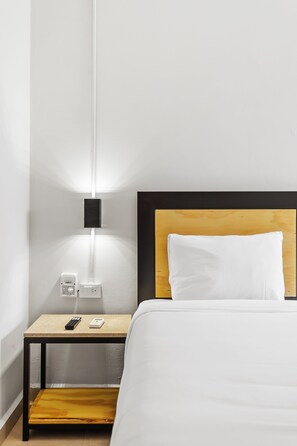 Habitación de hotel con cama, mesita de noche, control remoto y toma de corriente.