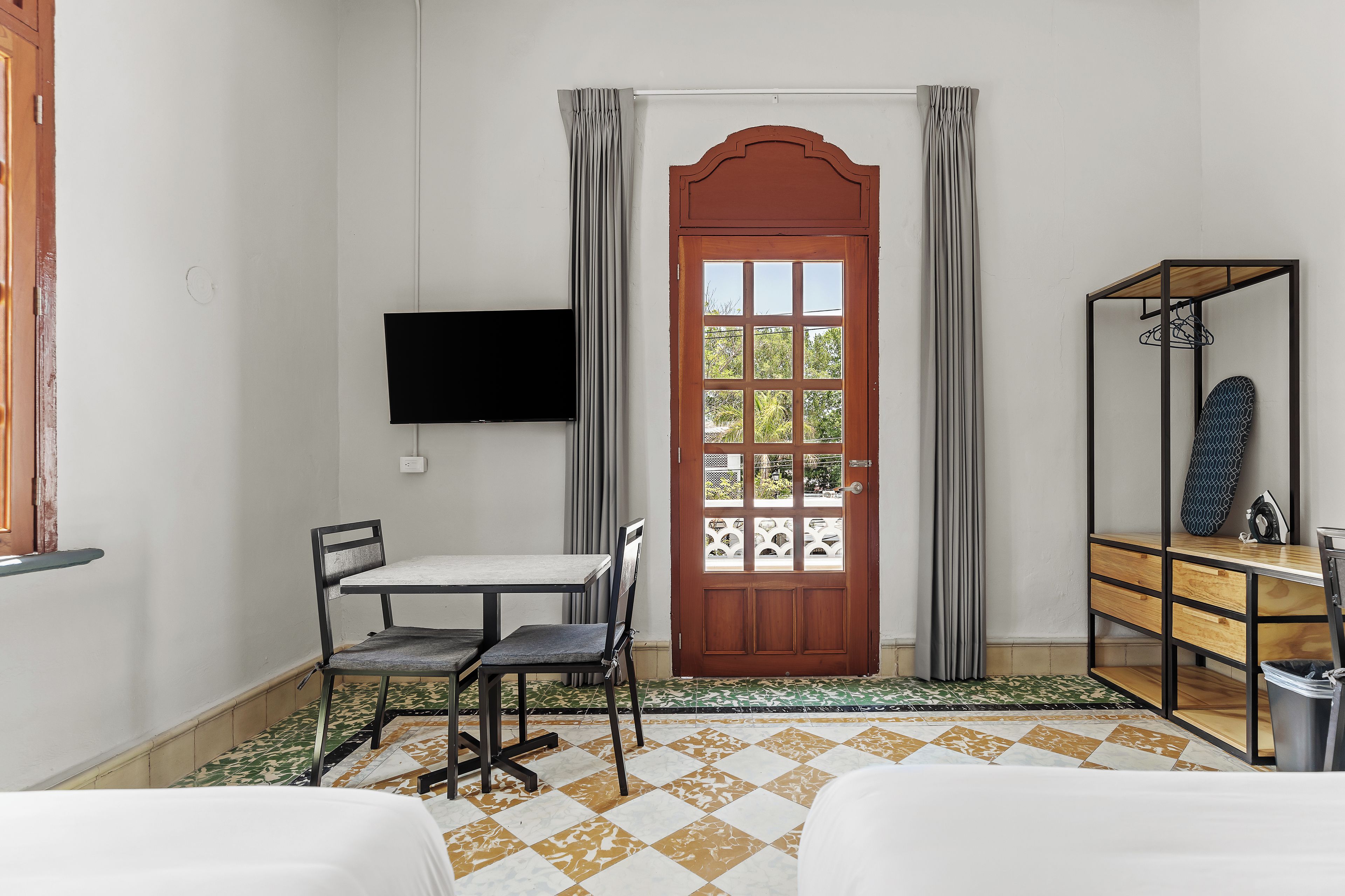 A room with a wooden door, a television mounted on the wall, a table with chairs, and a bed with a patterned bedspread.