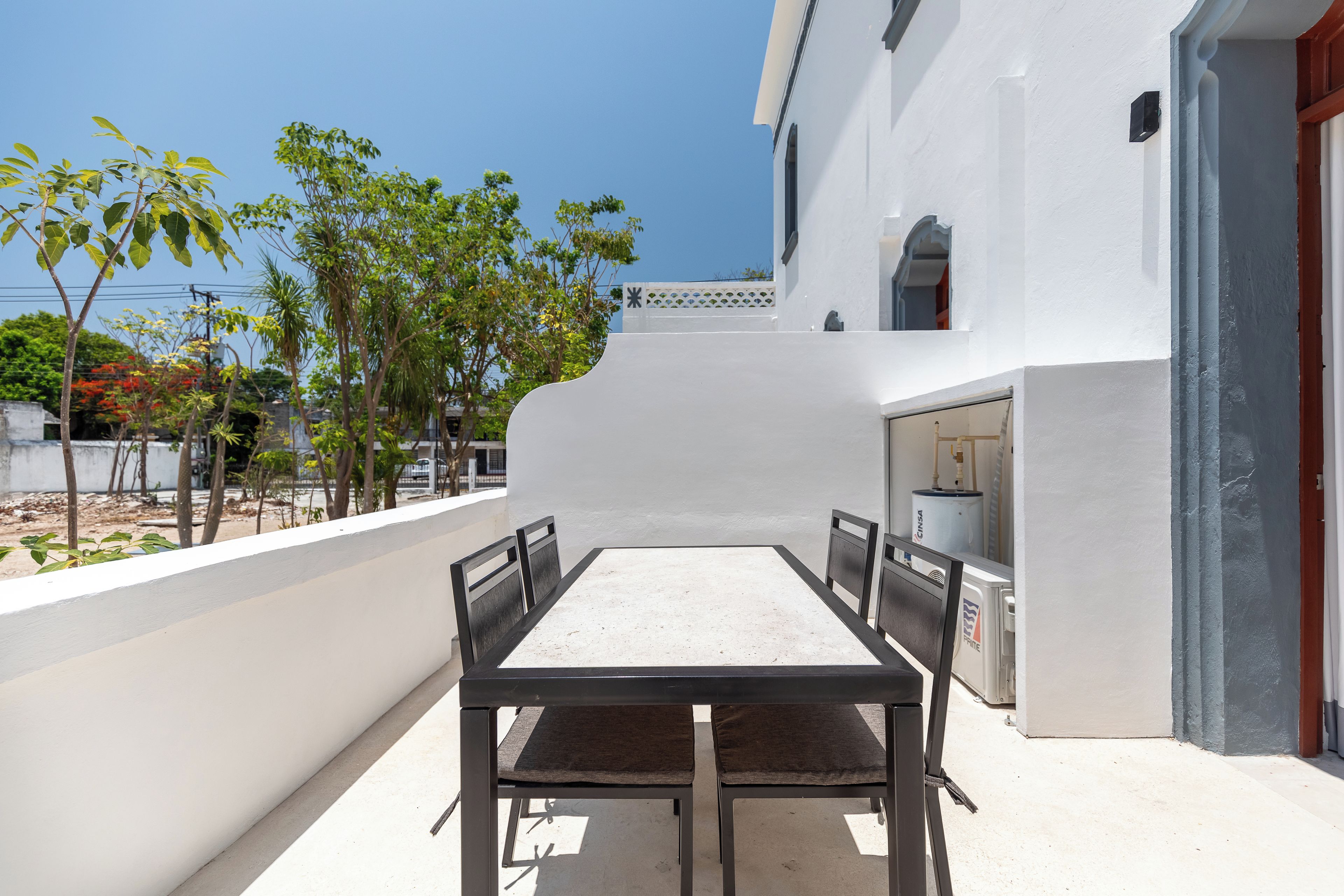 Eine Terrasse im Freien mit Tisch und Stühlen, einer weißen Wand und Blick auf Bäume sowie ein Gebäude im Hintergrund.