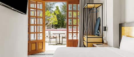 A hotel room with a large bed, a wooden door leading to an outdoor area, a small table with a chair, and a nightstand with an iron.