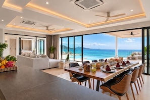 A modern hotel room with a dining area, large windows with a view of the sea, and a ceiling fan. - Sol Sofia 4BR/Seaview/Pool/BF/Staffs (Koh Samui)