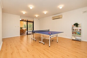 A room with a ping pong table, wooden flooring, a bookshelf with books and decorative items, and a door leading to an outdoor area. - Spacious 3-Bedroom Marion Home | Alfresco Living | Table Tennis (Marion)