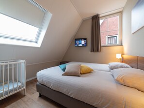 A bedroom with a sloped ceiling, a bed with pillows, a crib, a television on the wall, and a window with curtains. - Luxury villa with private wellness (Colijnsplaat)