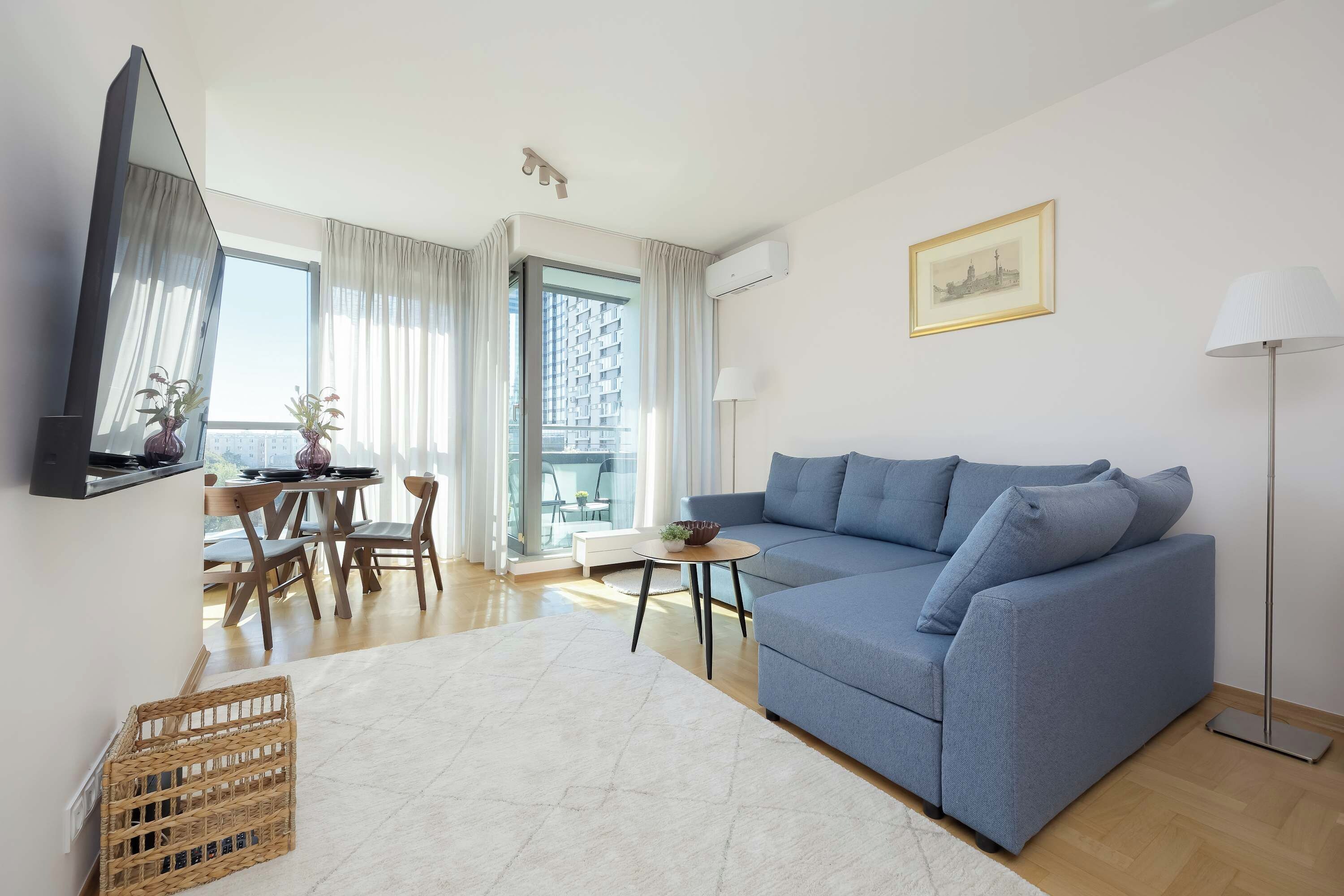 A modern living room with a blue sofa, a dining table with chairs, a TV mounted on the wall, and a large window with curtains.