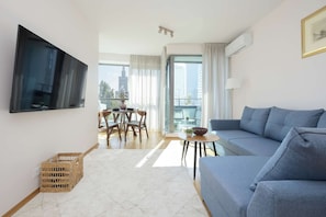 Una sala de estar moderna con un televisor de pantalla plana, un sofá, una mesa de comedor con sillas y vistas a la ciudad a través de amplios ventanales.