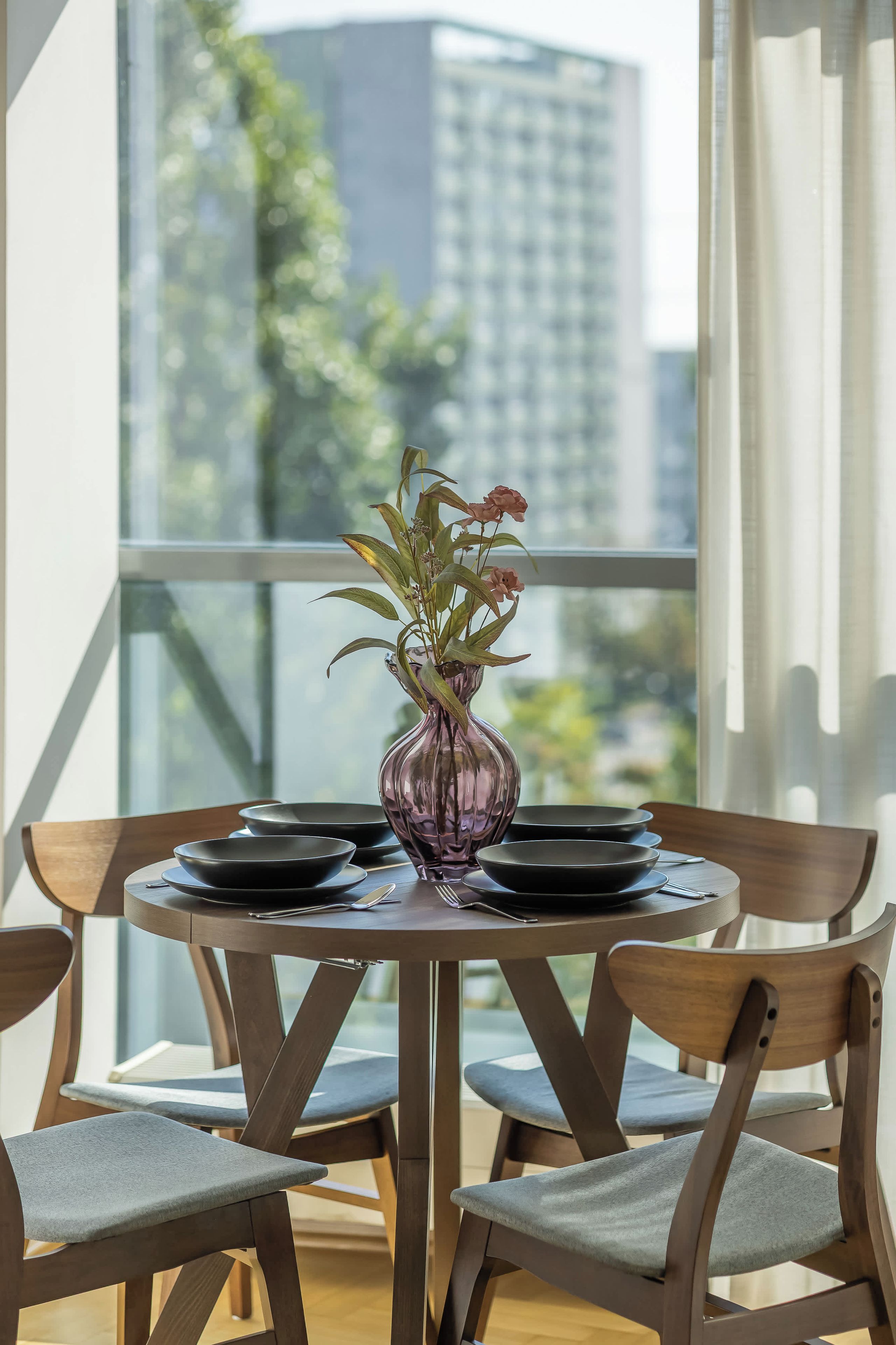 Una mesa de comedor con un jarrón de flores, platos negros y cubiertos, en una habitación con ventanas amplias y sillas de madera.