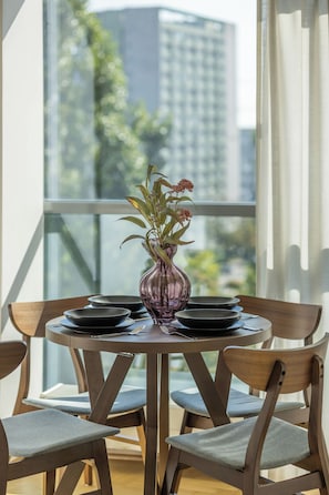 Una mesa de comedor con un jarrón de flores, platos negros y cubiertos, en una habitación con ventanas amplias y sillas de madera.